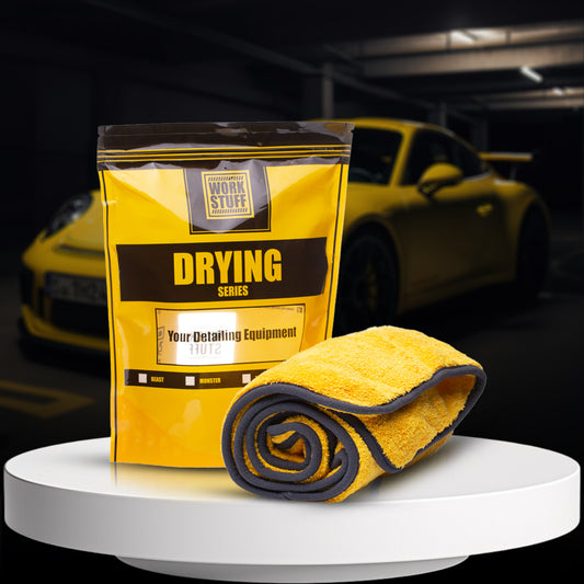A yellow Work Stuff Beast Drying Towel rolled up next to its packaging, which is also yellow with text and branding, placed on a white platform with a car in the background.