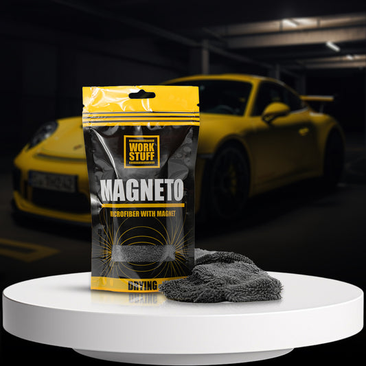 A packet of Work Stuff Magneto Microfiber placed on a white surface with a yellow car in the background. The microfiber appears gray and there are two of them, with integrated magnets visible on the packaging.