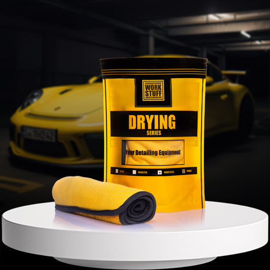 A rolled-up yellow microfiber drying towel displayed in front of its packaging, which has 'Drying' written on it, with a yellow sports car in the background.