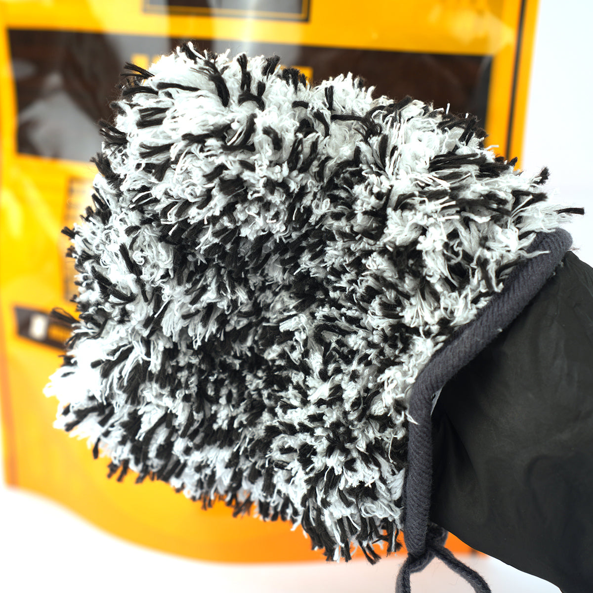 Black and white car cleaning mitt held by a gloved hand with a yellow car in the background.